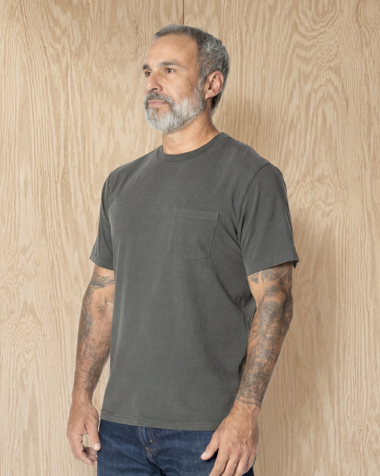 Man wearing a Steel Grey Pocket t-shirt and jeans against a wood background. Model is 5'11", 198lbs wearing size L