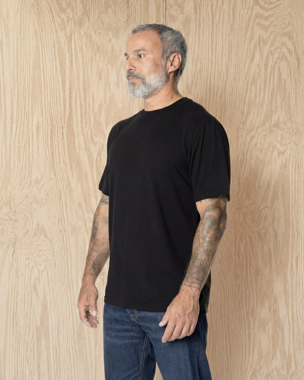 Man turned to the side, wearing a black Premium t-shirt against a wood background. Model is 5'11", 198lbs wearing size L