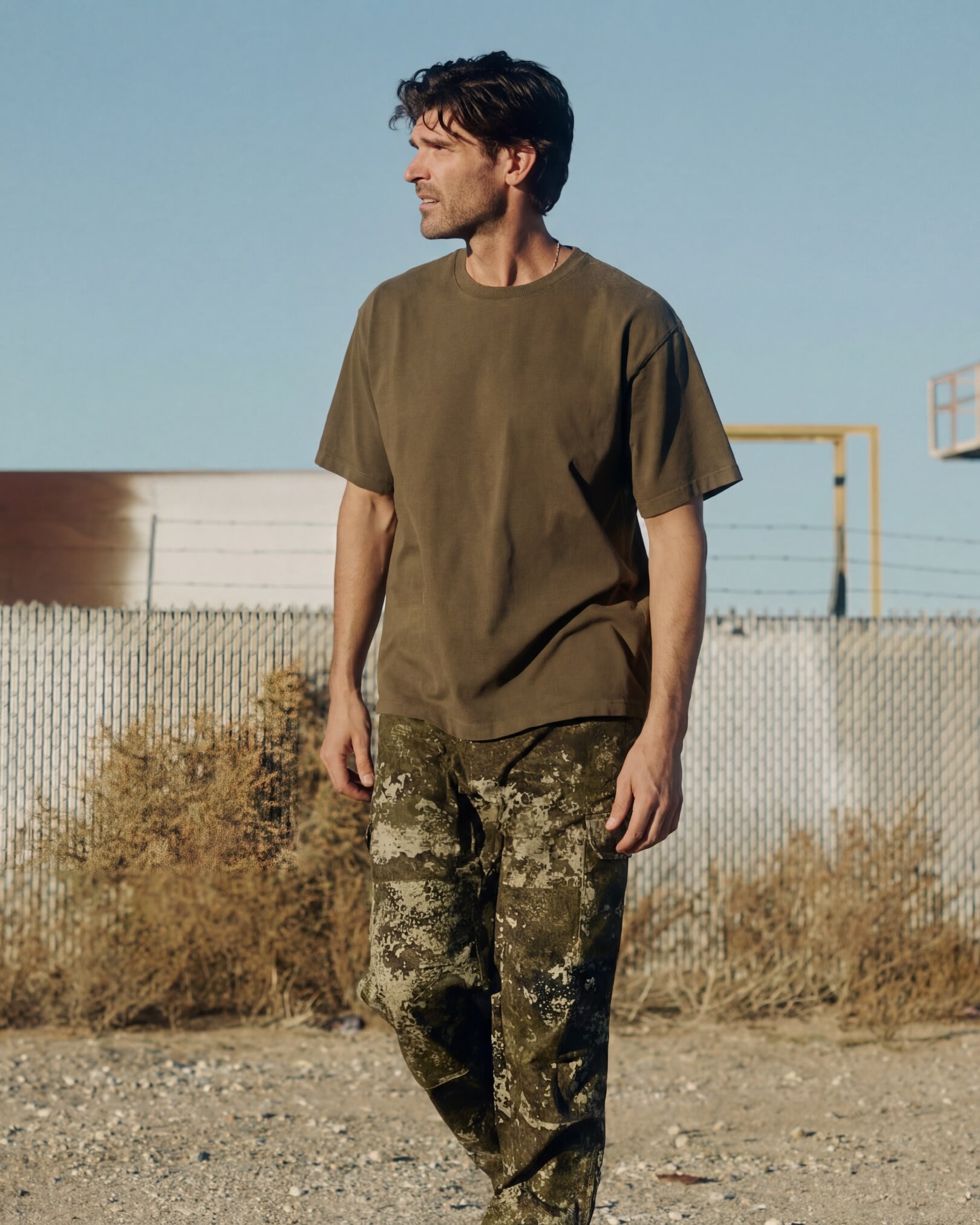 Man wearing a military green t-shirt and camouflage pants standing in an outdoor setting with a clear sky. Model is 6'1", 170lbs wearing size L