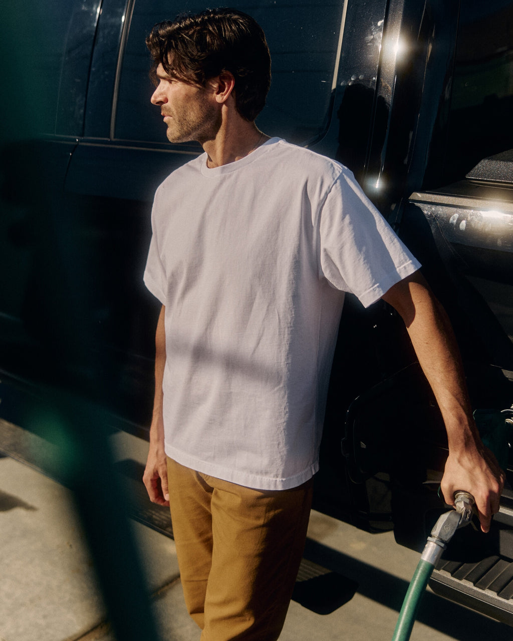 Man in a white t-shirt and brown pants standing next to a vehicle, holding a tool. Model is 6'1", 170lbs wearing size L
