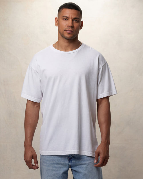 Man wearing a plain white drop shoulder t-shirt and light blue jeans against a beige background. Model is 6'0" wearing size L