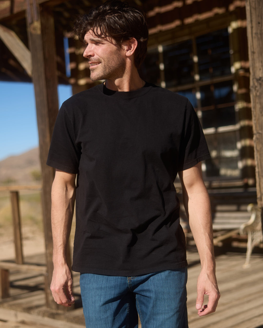 Man wearing a black t-shirt and blue jeans standing in front of a wooden structure. Model is 6'1", 170lbs wearing size L
